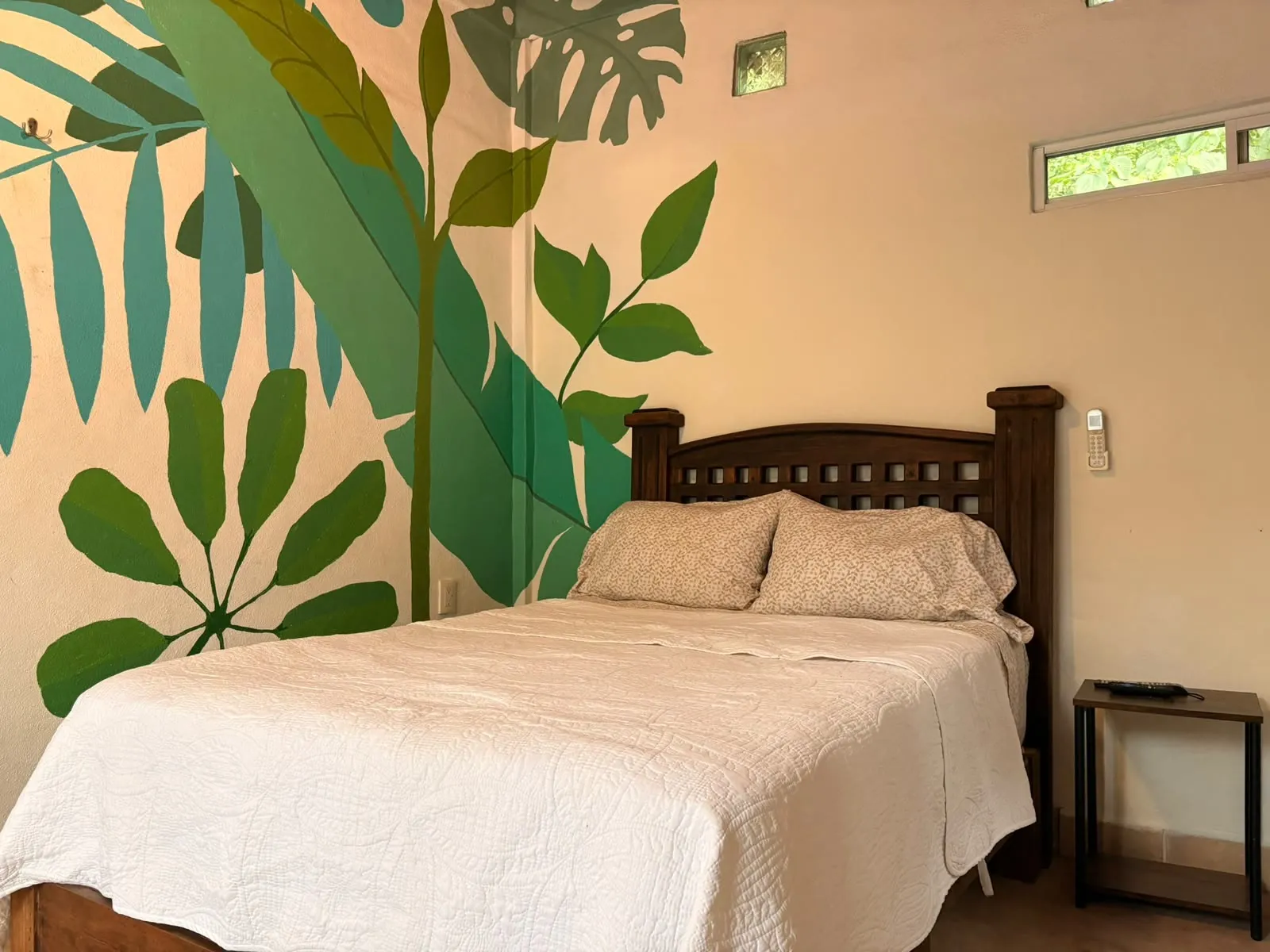 Cama vestida en blanco frente a un mural de palmas, con luz de la mañana.